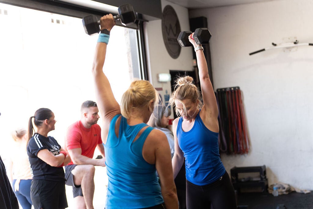 Two women in a gym lifting dumbbells above their head